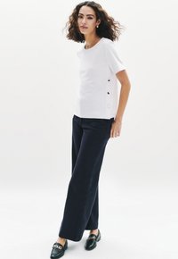 Top blanco de manga corta con cuello redondo, botones dorados en el costado. Combinado con pantalones amplios azul marino y mocasines negros con un adorno de cadena.