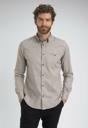 Homme aux cheveux bouclés et à la barbe portant une chemise beige à boutons avec une poche poitrine et un pantalon noir, se tenant avec les mains dans les poches.