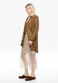 Camisa larga marrón y texturizada con capas, sobre una falda de beige, con longitud a la rodilla y plisada. Tela translúcida y fluida en el dobladillo de la falda. Zuecos marrones completan el atuendo.