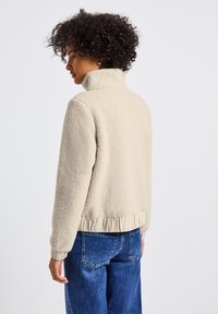 Giacca in fleece beige con colletto alto e orlo a coste. Tessuto morbido, vestibilità rilassata, abbinata a jeans in denim blu. Vista posteriore.