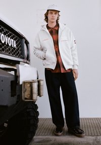 Šedá mikina na zip přes červenou kostkovanou košili, doplněná volnými tmavými kalhotami a černými botami. V pozadí je černé vozidlo Toyota.