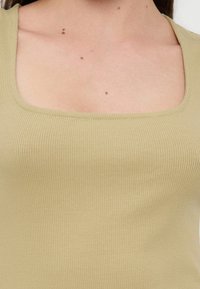 Κοντινό πλάνο στο πάνω μέρος του στήθους και το λαιμό ενός ατόμου που φοράει ριπτό, μπεζ τοπ με τετραγωνισμένη λαιμόκοψη και εμφανή σπίλους στο δέρμα.