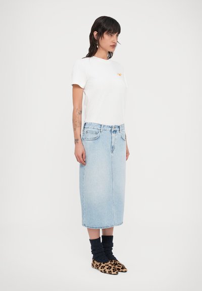 Λευκό βαμβακερό t-shirt με πορτοκαλί λογότυπο, συνδυασμένο με φούστα μίντι από ανοιχτό μπλε τζιν, σκούρες πλεκτές κάλτσες και παπούτσια με λεοπάρ σχέδιο.