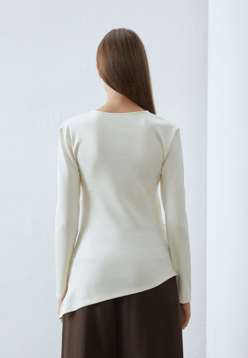 Długi rękaw z kremową bluzką o okrągłym dekolcie i zaokrąglonym dołem, wykonaną z gładkiego materiału, zestawioną z szerokimi brązowymi spodniami.