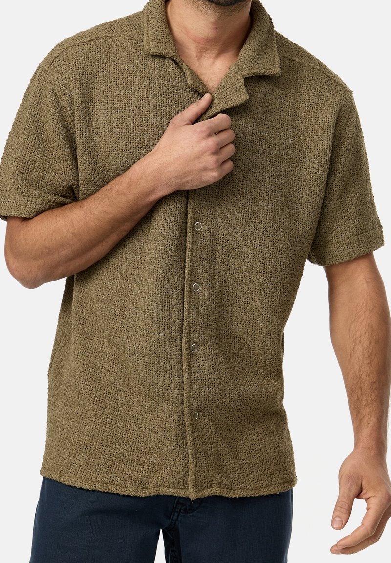 Homme portant une chemise texturée vert olive à manches courtes avec boutons et col évasé, ajustant le col à la main, associée à un pantalon sombre.