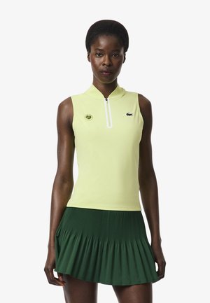 Donna che indossa un top smanicato giallo pallido con zip e loghi e gonna plissettata verde scuro, in piedi su sfondo bianco.