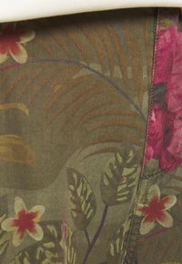 Gros plan d’un tissu avec un motif floral tropical dans des tons verts, marron et rouge avec des coutures visibles le long du bord.
