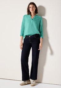 Camicia di lino turchese con scollo a V, abbinata a jeans dritti scuri e sneakers beige chiaro, esposta su uno sfondo neutro.