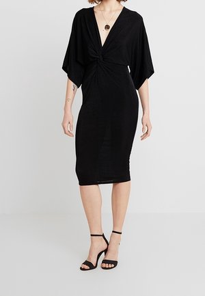 Kobieta ubrana w czarną sukienkę sięgającą kolan z głębokim dekoltem w kształcie litery V, z przodem związanym na supeł, szerokimi rękawami oraz czarne szpilki na kostkę z paskiem.