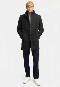 Abrigo de lana gris oscuro con cuello alto, diseño de un solo pecho y cierre de botones. Combina con pantalones azul marino y zapatillas blancas.