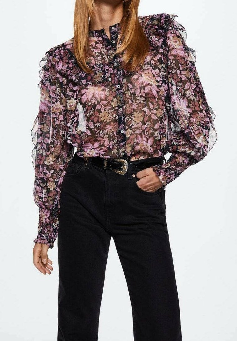 Femme portant un chemisier transparent à motif floral avec des détails à volants et des manches bouffantes, associé à un pantalon noir taille haute et une ceinture noire.