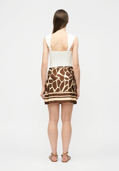 Γυναίκα που φαίνεται από πίσω, φορώντας λευκό τοπ χωρίς μανίκια, μίνι φούστα με καφέ και μπεζ μοτίβο καμηλοπάρδαλης και ταμπά σανδάλια, σε απλό φόντο.