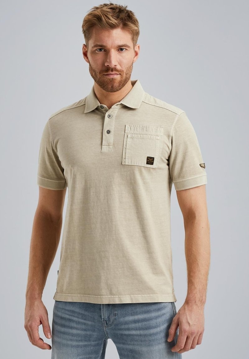 T-shirt polo beige en coton, avec un col à boutons, une poche sur la poitrine et un détail de logo subtil sur la manche. Manches courtes et coupe décontractée.
