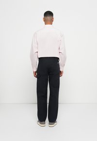 Mies, der ein hellrosa Langarmhemd, schwarze Cargo-Hosen und graue Turnschuhe trägt, steht entspannt mit den Händen an den Seiten vor einer weißen Wand.