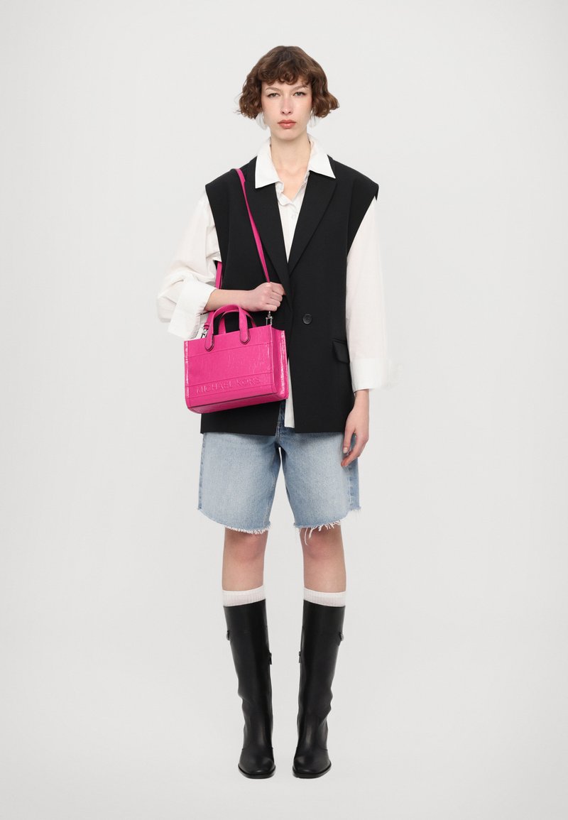 Borsa a tracolla con trama di coccodrillo rosa, dotata di manici doppi e tracolla, abbinata a un gilet nero, una camicia bianca, pantaloni shorts in denim e stivali neri.