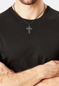 Uomo con le braccia incrociate, indossa una camicia nera e una collana con un pendente a forma di croce in fibra di carbonio attorno al collo.