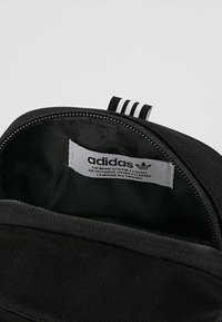 Černý batoh adidas uvnitř s zeleným podšívkou, obsahující textilní štítek a zipové zapínání. Tři bílé pruhy na vnější horní straně.