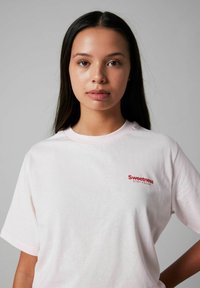 Lichtroze katoenen t-shirt met een ronde hals, korte mouwen en een rood logo met de tekst "Sweetness" en "STAY FRESH" op de borst.