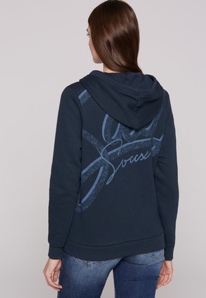 Dunkelblaues Kapuzensweatshirt aus weichem Material, mit einem strukturierten Grafikdesign, gerippten Bündchen und Saum, kombiniert mit Jeans.