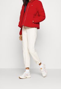 Giacca a vento rossa con cappuccio, chiusura a zip e due tasche frontali. Indossata sopra una maglietta blu navy, abbinata a pantaloni bianchi arrotolati e sneakers.