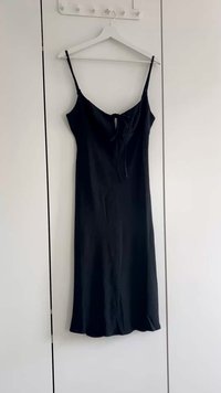 Schwarzes ärmelloses Kleid mit dünnen Trägern und einer vorderen Bindeschließe, aus glattem Stoff gefertigt, mit einem schwingenden Saum. Hängt an einem weißen Kleiderbügel.