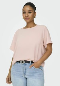 Różowa bluzka z krótkim rękawem o luźnym kroju i okrągłym dekoltem, zestawiona z jasnoniebieskimi dżinsami i czarnym paskiem z okrągłą srebrną klamrą.