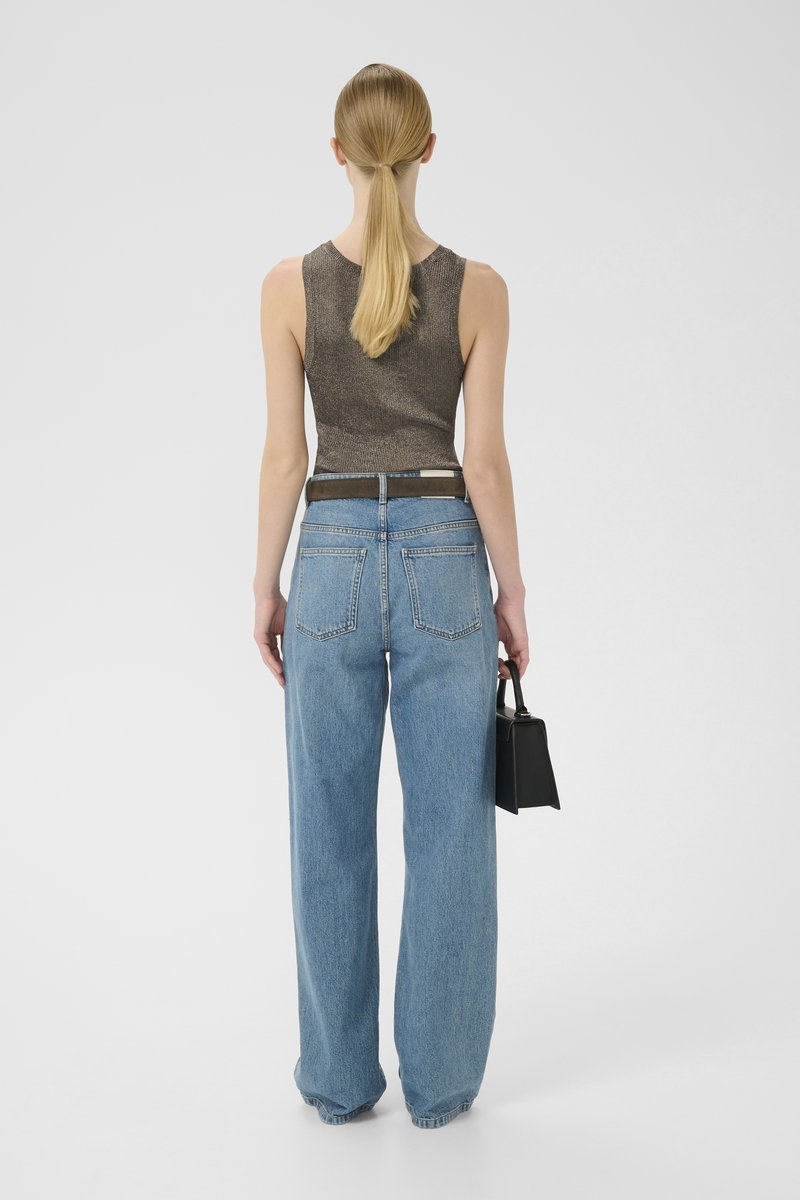 Woman with blonde ponytail wearing sleeveless brown top and wide-leg blue jeans, holding a small black handbag, standing against plain background.