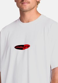 Hvid bomulds T-shirt med et rødt og sort oval logo med teksten "Billabong" i centrum. Korte ærmer, rund halsudskæring. Glat tekstur.