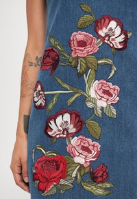 Denimbeklædning med detaljeret blomsterbroderi i nuancer af rød, pink og hvid, med grønne blade på en blå baggrund.