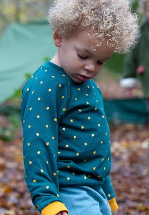 Türkisfarbenes Langarmshirt mit gelbem Sternenmuster, rundem Ausschnitt und gelben Bündchen, kombiniert mit hellblauen Cordhosen.
