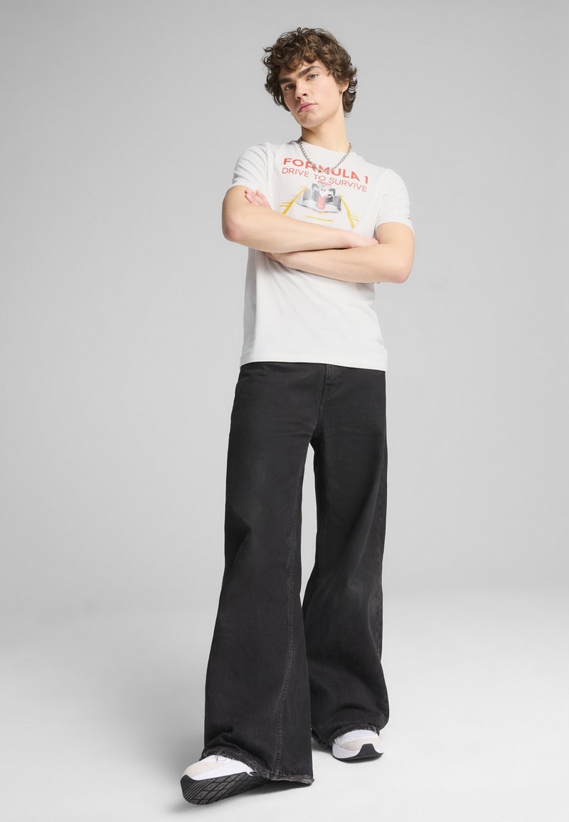 Hvid t-shirt med et rødt og gult "Formula 1" grafik, parret med vide sorte jeans og hvide sneakers med sorte såler.