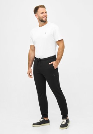Weißes T-Shirt mit kleinem Logo, kombiniert mit schwarzen Jogginghosen mit elastischem Bund und Bündchen, getragen mit schwarzen Sneakern.