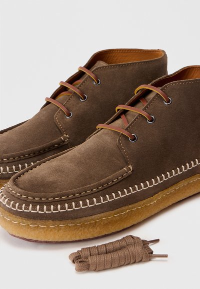 Chaussures chukka en daim marron avec des accents en cuir tan, des détails cousus, une semelle en caoutchouc et des lacets assortis reposant à côté.