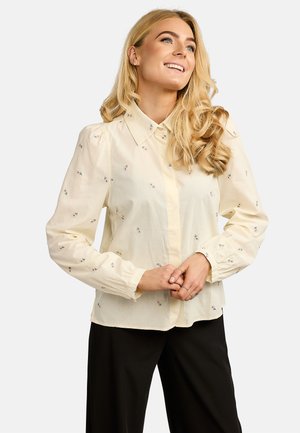 Blonde Frau lächelnd, trägt eine cremefarbene Langarmbluse mit kleinen Blumenmustern und schwarze Hose, die Hände vor der Taille verschränkt.