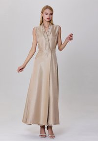 Robe maxi en satin beige avec un décolleté en V profond, sans manches, boutons décoratifs et fente sur le côté. Texture lisse et drapé élégant.