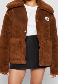 Bruine jas van namaakbont met een knoopsluiting, grote zakken en een kraag; heeft een logopatch op de linkerborst; met een gestructureerd oppervlak.