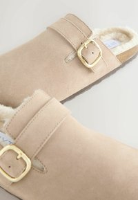 Zuecos de ante beige con un suave forro interior, que cuentan con una hebilla de metal dorado y un diseño de dos correas. Suela de goma texturizada para mayor agarre.