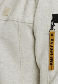 Sweat à capuche gris clair avec texture matelassée, comportant une étiquette beige et une étiquette en tissu jaune avec du texte noir fixée par un petit clip.
