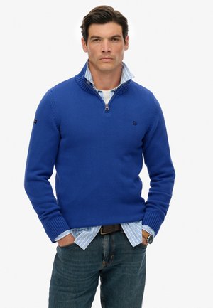 Königsblauer Strickpullover mit hohem Kragen, Reißverschluss, gerippten Bündchen und einem Logo auf der Brust, getragen über einem hellblau gestreiften Hemd.