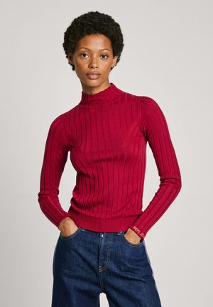 Maglione a collo alto rosso a coste con maniche lunghe, realizzato in un materiale morbido. Presenta una vestibilità aderente e un logo sul polsino. Abbinato a jeans in denim.