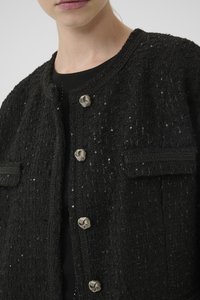 Černé tvídové sako s texturovaným povrchem, stříbrnými kulatými knoflíky a malými bočními kapsami. Má kulatý výstřih a složité prošívání.