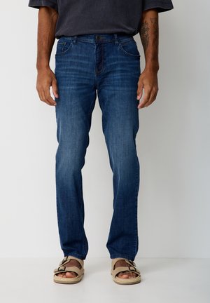 Man wearing blue denim jeans and beige sandals, standing against a white background with tattoo visible on left forearm.