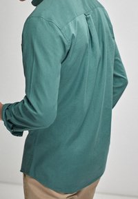 Maglietta a maniche lunghe in teal con vestibilità relaxed, dotata di una piega sul retro, polsini con bottoni e orlo curvo. Tessuto morbido, design minimalista.