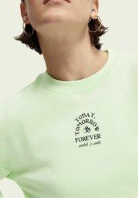 Felpa verde menta con scollo rotondo. Design testuale nero con la scritta "OGGI, DOMANI & PER SEMPRE" e "scotch & soda" in un font stilizzato.