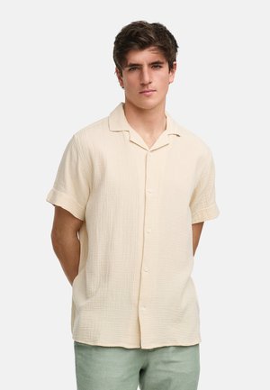 Hellbeige Hemd mit kurzen Ärmeln aus strukturiertem Stoff, Knopfverschluss und lockerer Kragenform. Kombination mit hellgrünen Hosen.