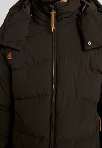 Mørkegrøn puffjakke med høj krave, quiltet design, frontlynlås med brun snor og læderdetaljer.