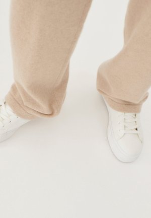 Person trägt beige Hose und weiße Schnürsneaker, steht auf einer weißen Oberfläche.