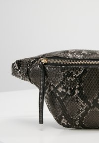 Sac banane à motif peau de serpent noir et gris avec fermeture éclair dorée et tirette en cuir sur un fond clair uni.
