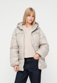 Chaqueta de plumas gris claro con capucha, con un diseño acolchado, cremallera frontal y bolsillos laterales, llevada sobre una camisa de botones beige.