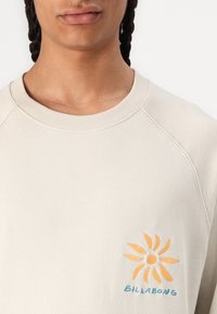 Hellbeige Sweatshirt mit Rundhalsausschnitt und Raglanärmeln. Es hat ein gesticktes Sonnenlogo in Orange und Blau auf der Brust.